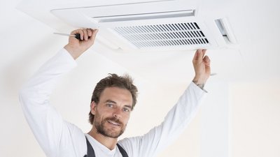 Ein Handwerker installiert eine Klimaanlage an der Decke und hält dabei einen Schraubendreher in der Hand.